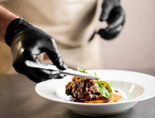Kokk med svarte hansker legger gourmetmat på en hvit tallerken