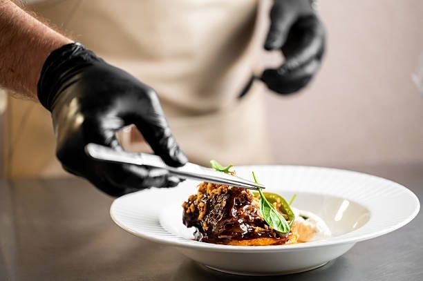 Kokk med svarte hansker legger gourmetmat på en hvit tallerken